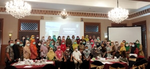 A large group of people are gathered in a conference room for a 'Sensitivity Training' session. Most attendees are wearing face masks. The room is decorated with chandeliers and a large screen displaying the title of the seminar. Attendees are dressed in various colorful garments.