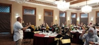 A group of people are seated around tables in a well-decorated room with chandeliers and wooden accents. Many are raising their hands, indicating an interactive session or discussion. Most attendees are wearing masks, and a speaker at the front appears to be leading the session.