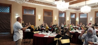 A group of people are seated around tables in a well-decorated room with chandeliers and wooden accents. Many are raising their hands, indicating an interactive session or discussion. Most attendees are wearing masks, and a speaker at the front appears to be leading the session.