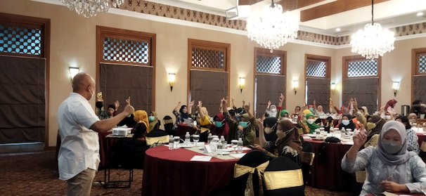 A group of people are seated around tables in a well-decorated room with chandeliers and wooden accents. Many are raising their hands, indicating an interactive session or discussion. Most attendees are wearing masks, and a speaker at the front appears to be leading the session.
