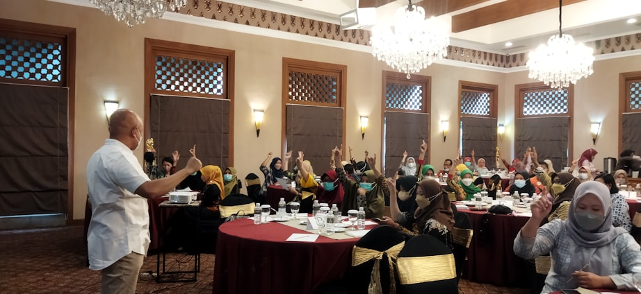 A group of people are seated around tables in a well-decorated room with chandeliers and wooden accents. Many are raising their hands, indicating an interactive session or discussion. Most attendees are wearing masks, and a speaker at the front appears to be leading the session.