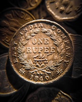 A close-up view of an antique coin from the East India Company, with intricate engravings including the words 'ONE RUPEE' and the year '1840'. The coin is surrounded by a detailed laurel wreath pattern.