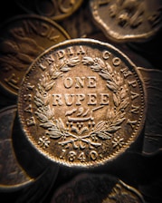 A close-up view of an antique coin from the East India Company, with intricate engravings including the words 'ONE RUPEE' and the year '1840'. The coin is surrounded by a detailed laurel wreath pattern.