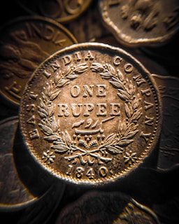 A close-up view of an antique coin from the East India Company, with intricate engravings including the words 'ONE RUPEE' and the year '1840'. The coin is surrounded by a detailed laurel wreath pattern.