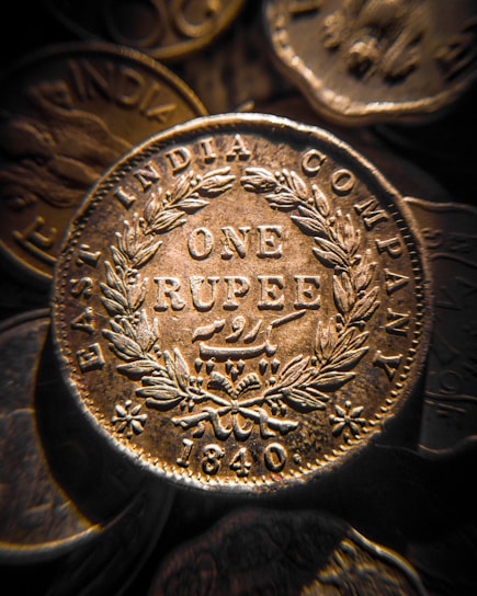 A close-up view of an antique coin from the East India Company, with intricate engravings including the words 'ONE RUPEE' and the year '1840'. The coin is surrounded by a detailed laurel wreath pattern.