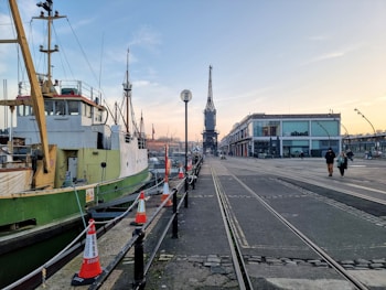 A dockside scene featuring a moored ship with a green and white hull alongside a pier. In the distance, a large industrial crane stands tall, adjacent to a modern building with the name 'shed' displayed on it. Several orange traffic cones line the walkway beside the ship, and two pedestrians are walking near the building. The atmosphere suggests early morning or late afternoon with soft lighting.