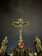 A crucifix stands prominently against a dark, cloudy sky. Flanking the crucifix are two statues in robes, depicting solemn figures. In front of the cross, a red glass lantern hangs, casting a subtle glow. Gold accents on the cross add contrast to the otherwise muted color palette.