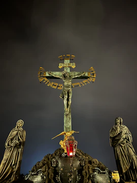 A crucifix stands prominently against a dark, cloudy sky. Flanking the crucifix are two statues in robes, depicting solemn figures. In front of the cross, a red glass lantern hangs, casting a subtle glow. Gold accents on the cross add contrast to the otherwise muted color palette.