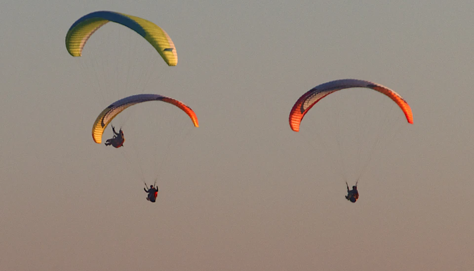 Three paragliders soar through clear blue sky with vibrant colored parachutes attached