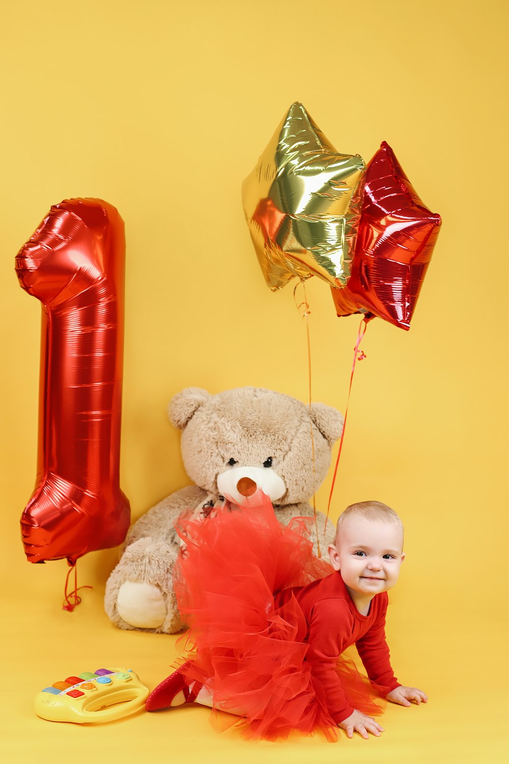 Un bebé sentado junto a un oso de peluche y globos