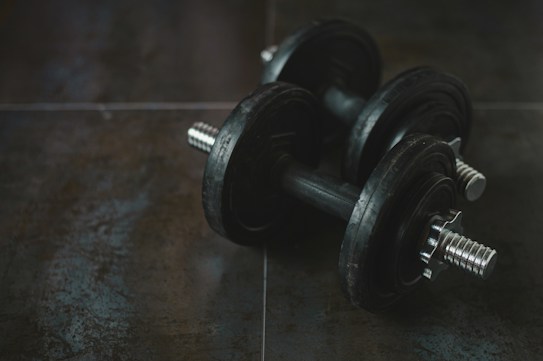 A pair of black dumbbells placed on a dark textured surface. The weights have a shiny metallic grip, contrasting with the matte finish of the weights themselves. The environment suggests a gym or workout setting.