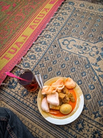 A dish featuring a variety of foods including crackers, boiled eggs, and chunks of meat or tofu in a red-orange sauce is placed on a patterned mat. Beside the plate sits a glass of dark-colored beverage with a pink spoon. The patterns on the mat include intricate designs in blue, beige, red, and yellow.