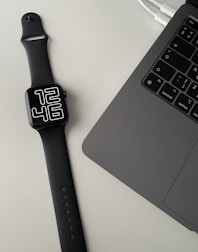 A black smartwatch with a digital clock face displaying 12:46 is placed on a light-colored surface next to a laptop. The laptop is open, showing a partial view of the keyboard and part of the touchpad. Two cables are also visible connected to the laptop.