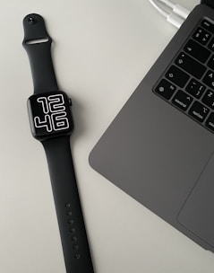 A black smartwatch with a digital clock face displaying 12:46 is placed on a light-colored surface next to a laptop. The laptop is open, showing a partial view of the keyboard and part of the touchpad. Two cables are also visible connected to the laptop.