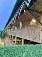 An open-air bamboo structure with a slanted roof, featuring decorative hanging lights and green accents. Below the lights, there are wooden tables and benches, suggesting a dining or communal area.