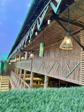 An open-air bamboo structure with a slanted roof, featuring decorative hanging lights and green accents. Below the lights, there are wooden tables and benches, suggesting a dining or communal area.