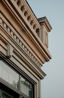 Architectural details of a classical building facade with ornate plasterwork. The design features decorative columns, structural intricacies, and a window with reflective glass. The plasterwork includes repetitive elements such as balusters and moldings in a light beige color, set against a clear sky.