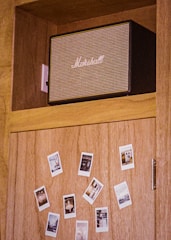 A wooden cabinet features a black and beige Marshall speaker on the top shelf. Below, several Polaroid photos are scattered across the cabinet door, affixed in a casual pattern.