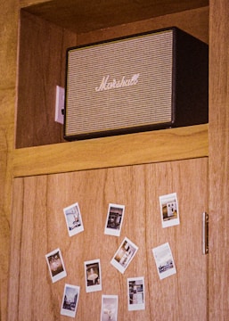 A wooden cabinet features a black and beige Marshall speaker on the top shelf. Below, several Polaroid photos are scattered across the cabinet door, affixed in a casual pattern.