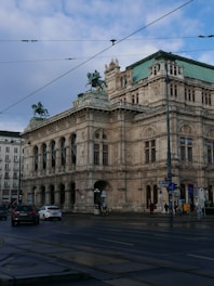A grand, historic building with ornate architectural details, featuring statues of horsemen on the roof. The structure appears to be a prominent cultural or civic building, possibly an opera house or museum. The street in front is lightly trafficked with cars and a few pedestrians visible.