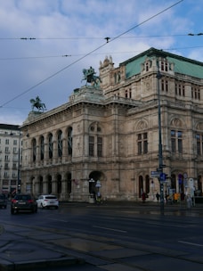 A grand, historic building with ornate architectural details, featuring statues of horsemen on the roof. The structure appears to be a prominent cultural or civic building, possibly an opera house or museum. The street in front is lightly trafficked with cars and a few pedestrians visible.
