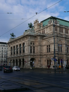A grand, historic building with ornate architectural details, featuring statues of horsemen on the roof. The structure appears to be a prominent cultural or civic building, possibly an opera house or museum. The street in front is lightly trafficked with cars and a few pedestrians visible.