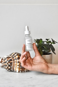 A hand holding a dropper bottle of skincare product labeled 'The Ordinary' with a focus on the bottle. In the background, there is a large decorative seashell and a small potted plant.
