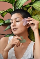 A person with short hair applies skincare with a gua sha tool to their cheek while sitting in front of large, vibrant green plants. The person's eyes are closed, and they wear a white tank top with lace detailing.