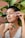 A person with short hair applies skincare with a gua sha tool to their cheek while sitting in front of large, vibrant green plants. The person's eyes are closed, and they wear a white tank top with lace detailing.