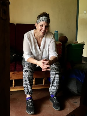 A person is sitting on a wooden bench with a relaxed posture, smiling warmly. The individual is dressed casually in patterned leggings, a white shirt, and sneakers. They are adorned with a headband and bracelets. The background includes a soft drink bottle, a green container, and other items, suggesting a cozy indoor setting.