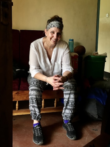A person is sitting on a wooden bench with a relaxed posture, smiling warmly. The individual is dressed casually in patterned leggings, a white shirt, and sneakers. They are adorned with a headband and bracelets. The background includes a soft drink bottle, a green container, and other items, suggesting a cozy indoor setting.