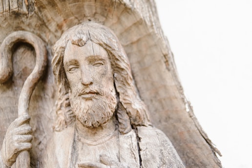 A detailed wooden carving of a bearded man with long hair, holding a shepherd's staff. The carving is integrated into the natural texture of the wood, giving it an organic and rustic appearance. The face of the man shows a calm and serene expression.