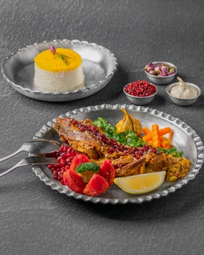 An elaborate meal set on a metallic tray, showcasing a spiced fish garnished with red berries and herbs, accompanied by vegetables such as tomatoes, carrots, and greens. Beside it, a small metallic bowl of saffron rice topped with a piece of saffron, and several smaller bowls containing ingredients like garlic, pomegranate seeds, and dried flowers are visible.