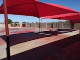 A shaded outdoor area with large red fabric canopies suspended over a concrete surface. The ground is divided into sections of red and gray tiles. The background features a tall wall with barbed wire, some trees, and simple structures.
