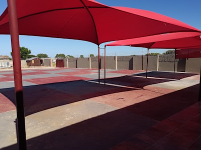 A shaded outdoor area with large red fabric canopies suspended over a concrete surface. The ground is divided into sections of red and gray tiles. The background features a tall wall with barbed wire, some trees, and simple structures.