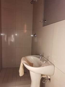 A bathroom features a white pedestal sink with silver faucets. A beige towel hangs over the edge of the sink. The walls are covered with large beige tiles, and a shower head is visible above the sink area. The lighting is soft, creating a calm and clean atmosphere.