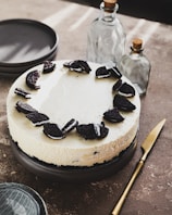 A cheesecake topped with crushed cookies is placed on a dark plate. Surrounding the plate are stacked gray dishes, a gold knife, and two glass bottles with cork stoppers. The scene is set on a textured brown surface with soft lighting creating subtle shadows.