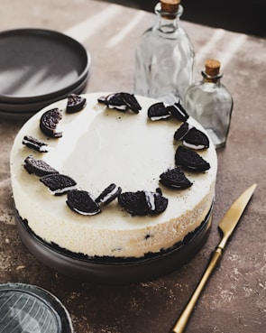 A cheesecake topped with crushed cookies is placed on a dark plate. Surrounding the plate are stacked gray dishes, a gold knife, and two glass bottles with cork stoppers. The scene is set on a textured brown surface with soft lighting creating subtle shadows.