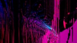 An abstract digital artwork featuring vibrant streaks and splashes of pink, blue, and purple against a predominantly black background. The pattern resembles a chaotic yet structured accumulation of vertical lines and scattered pixels, creating a vivid sense of movement and depth.