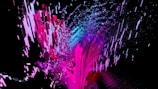 An abstract digital artwork featuring dynamic streaks and blocks of bright colors, predominantly pink and blue, emerging against a black background. The composition gives a sense of movement and fragmentation, with pixel-like patterns creating a vibrant and energetic visual effect.