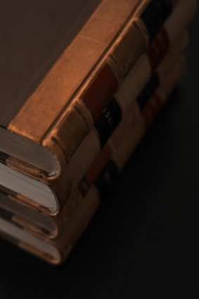 A stack of four vintage hardcover books with brown leather bindings and visible wear. The spines feature black and red labels with faint text. The books are positioned horizontally against a dark background, creating a classic and antique atmosphere.