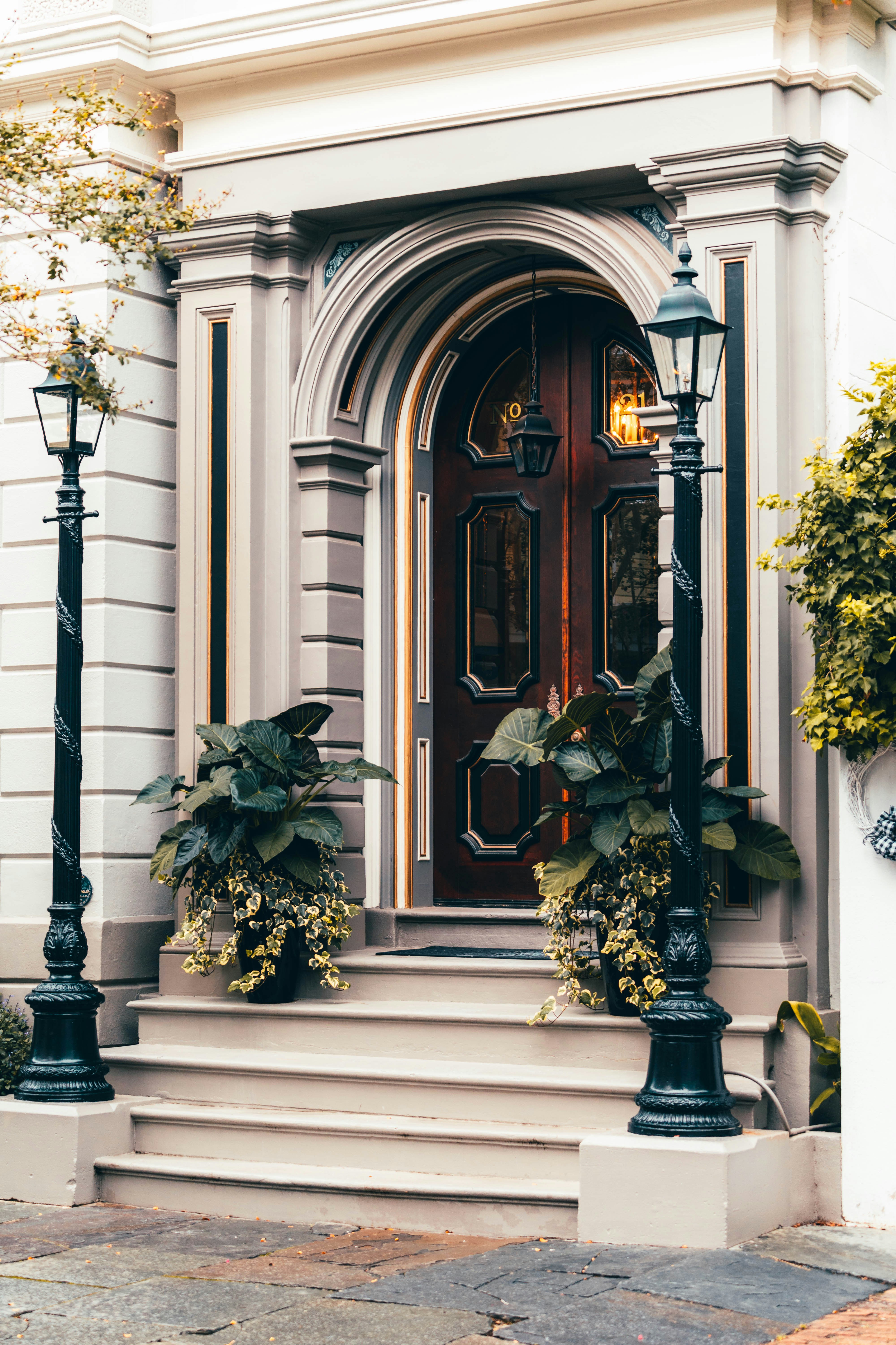 A classic wooden door with intricate panel designs and decorative molding is flanked by two black metal lampposts. The entrance is accentuated by potted plants with lush green leaves and cascading yellow flowers. The building facade features elegant architectural details in neutral tones, conveying a timeless sense of style.