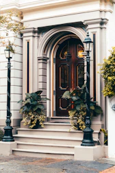 A classic wooden door with intricate panel designs and decorative molding is flanked by two black metal lampposts. The entrance is accentuated by potted plants with lush green leaves and cascading yellow flowers. The building facade features elegant architectural details in neutral tones, conveying a timeless sense of style.