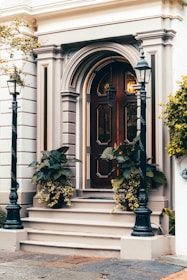 A classic wooden door with intricate panel designs and decorative molding is flanked by two black metal lampposts. The entrance is accentuated by potted plants with lush green leaves and cascading yellow flowers. The building facade features elegant architectural details in neutral tones, conveying a timeless sense of style.