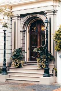 A classic wooden door with intricate panel designs and decorative molding is flanked by two black metal lampposts. The entrance is accentuated by potted plants with lush green leaves and cascading yellow flowers. The building facade features elegant architectural details in neutral tones, conveying a timeless sense of style.