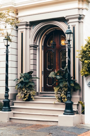 A classic wooden door with intricate panel designs and decorative molding is flanked by two black metal lampposts. The entrance is accentuated by potted plants with lush green leaves and cascading yellow flowers. The building facade features elegant architectural details in neutral tones, conveying a timeless sense of style.