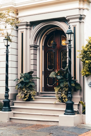 A classic wooden door with intricate panel designs and decorative molding is flanked by two black metal lampposts. The entrance is accentuated by potted plants with lush green leaves and cascading yellow flowers. The building facade features elegant architectural details in neutral tones, conveying a timeless sense of style.