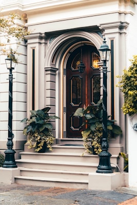 A classic wooden door with intricate panel designs and decorative molding is flanked by two black metal lampposts. The entrance is accentuated by potted plants with lush green leaves and cascading yellow flowers. The building facade features elegant architectural details in neutral tones, conveying a timeless sense of style.