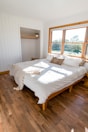 A bright and airy bedroom features a large double bed with white bedding and a cozy throw blanket. The room has wooden floors and white paneled walls. There is a small open closet with a hanging rod and a potted plant on a shelf. Large windows allow sunlight to flood into the room, offering a view of greenery outside.