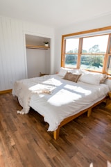 A bright and airy bedroom features a large double bed with white bedding and a cozy throw blanket. The room has wooden floors and white paneled walls. There is a small open closet with a hanging rod and a potted plant on a shelf. Large windows allow sunlight to flood into the room, offering a view of greenery outside.
